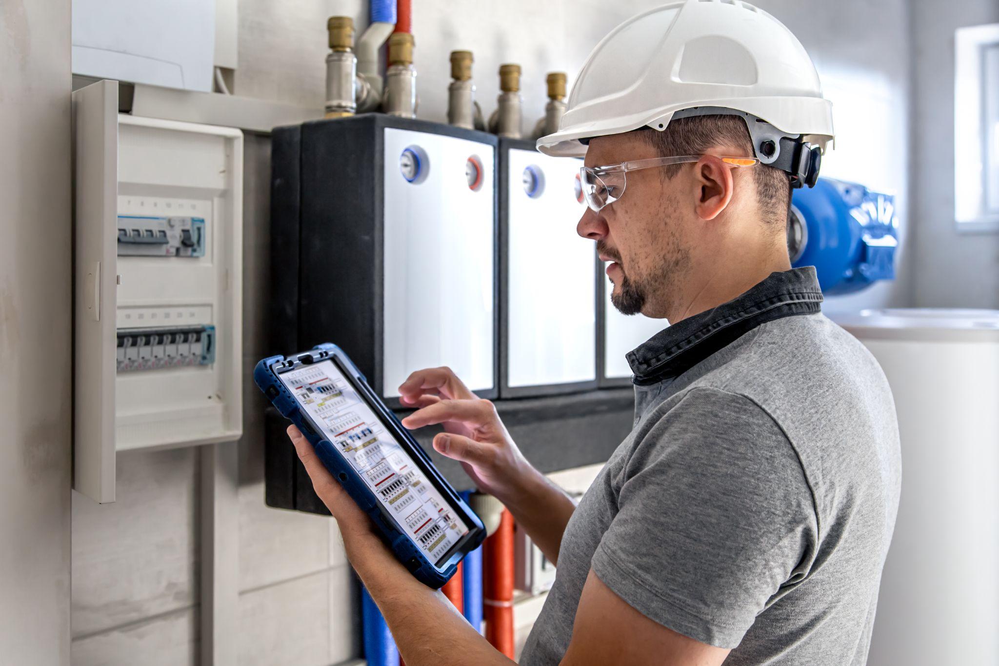 A technician in a hard hat checking a tablet beside a home battery and hot water system
