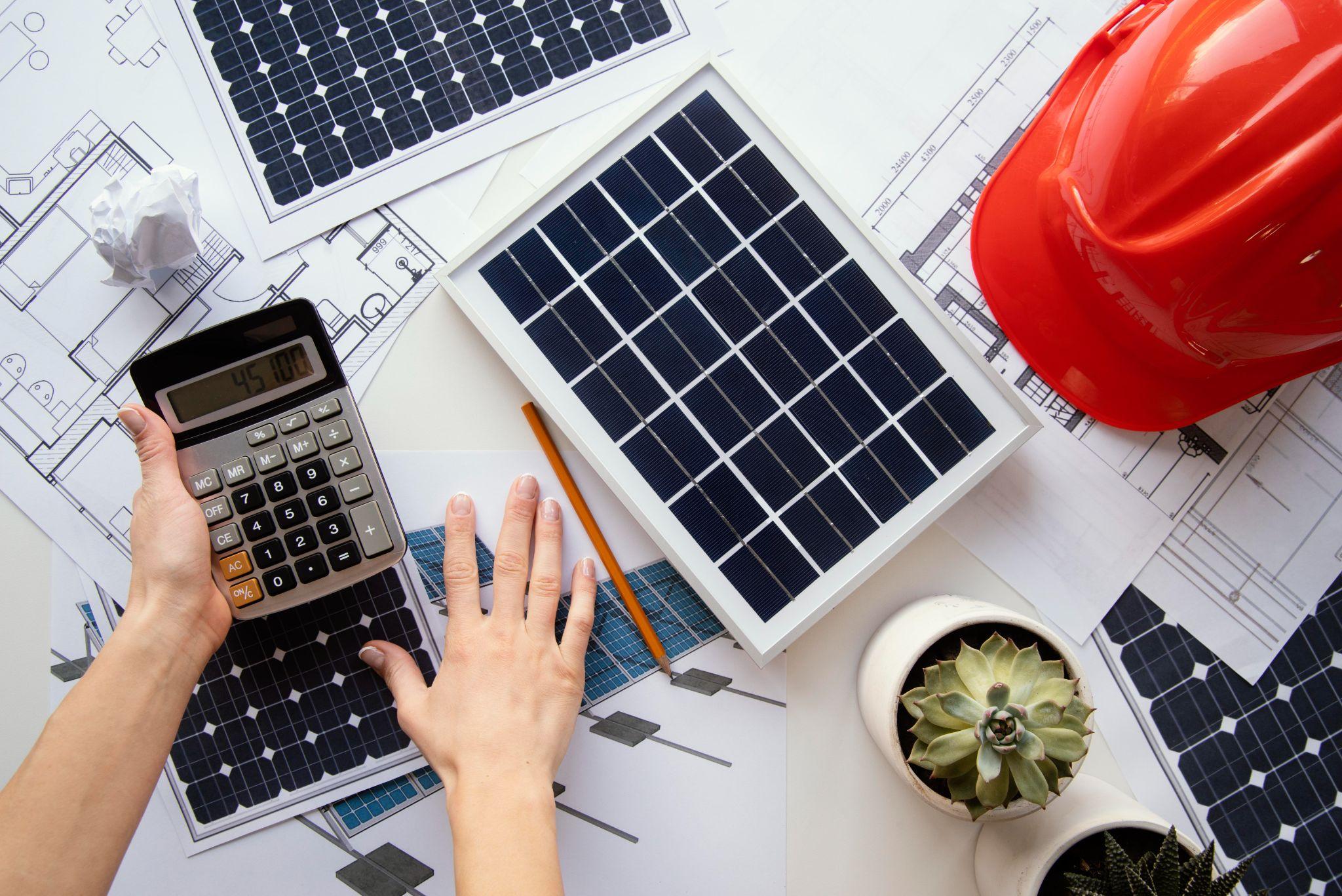A calculator and a small solar panel resting on building plans next to a red hard hat