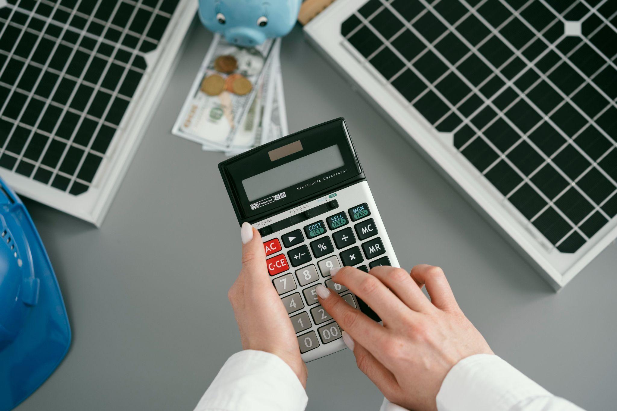 A person using a calculator at a desk with solar panels, banknotes and a coin-filled piggy bank