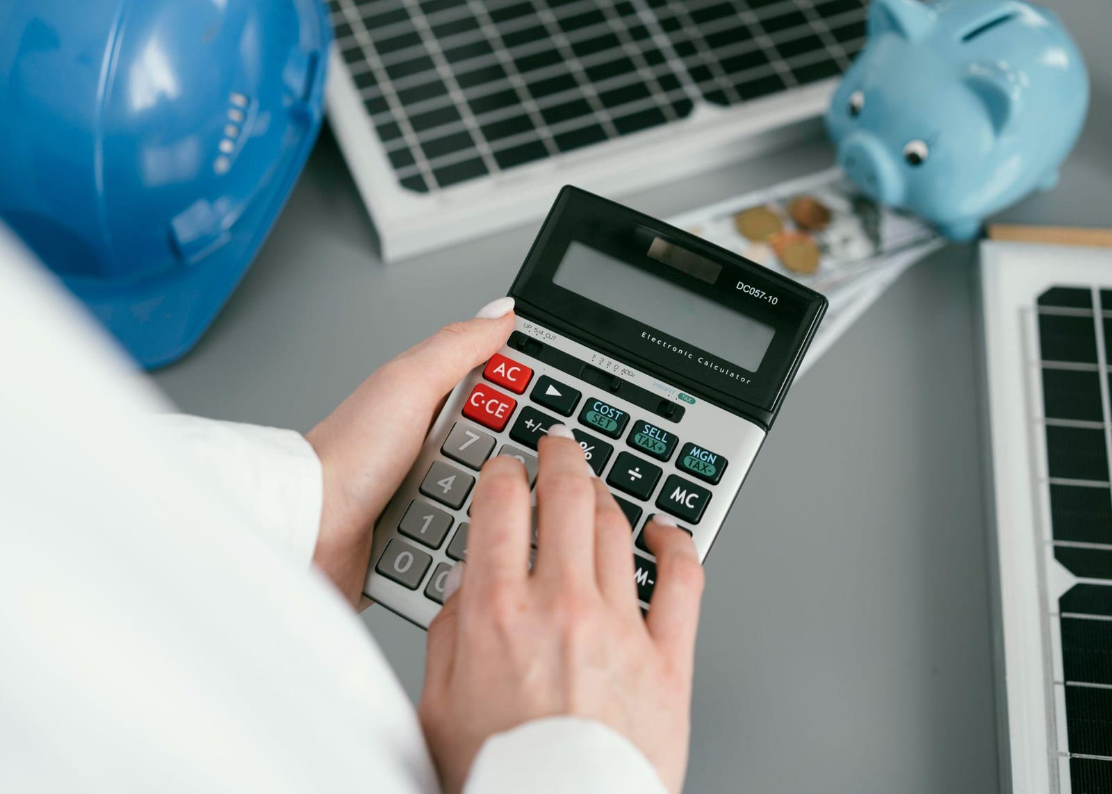 A person using a calculator beside a solar panel, a blue hard hat and a piggy bank